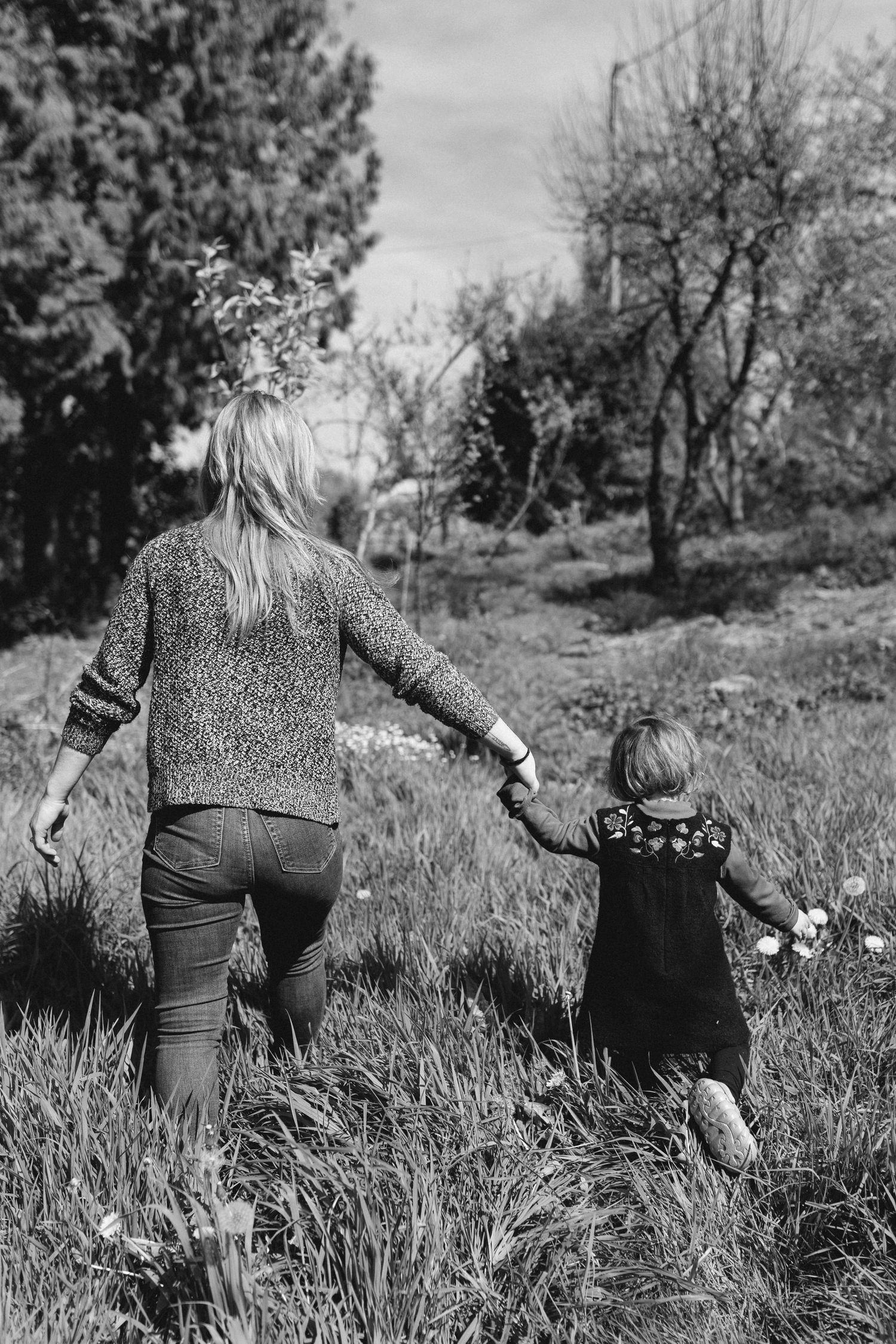 Vue arrière en noir et blanc d'une mère et de son jeune enfant se tenant la main et marchant dans un champ herbeux bordé d'arbres.