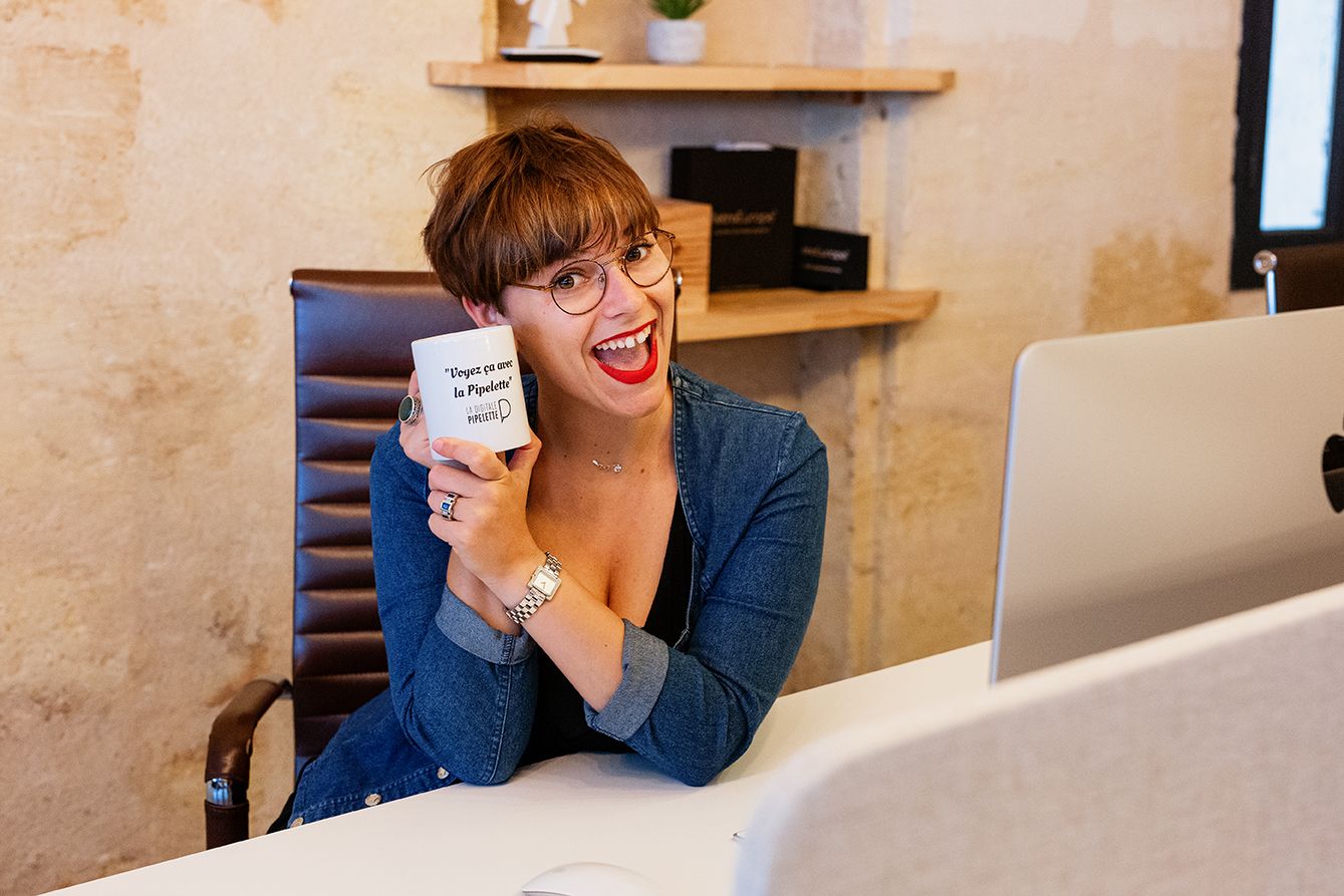Smiling woman holding a white mug laughing at her desk in a bright, modern workspace.