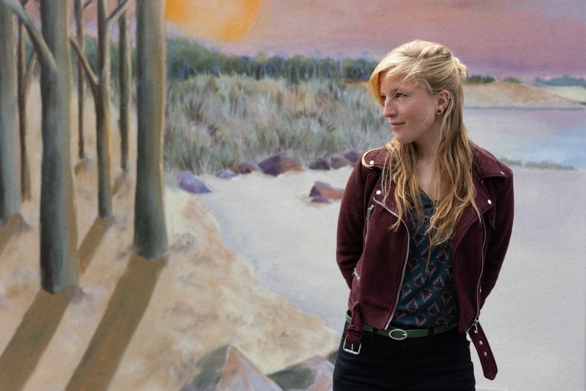 Blonde woman in burgundy leather jacket standing before a coastal landscape painting in an art gallery.