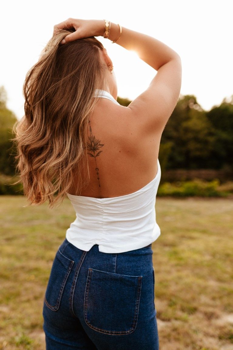 Femme en débardeur blanc dos nu et jean, dévoilant un tatouage dans le dos, posant dans un champ ensoleillé.