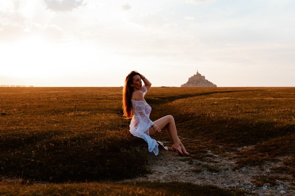 Woman in white dress sitting on path at sunset with Mont Saint-Michel in background.