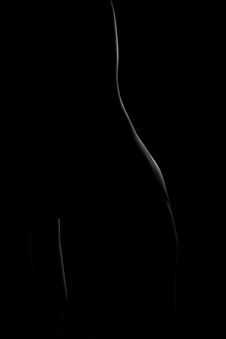 Black and white fine art silhouette photograph highlighting the curves of a human torso against a dark background.