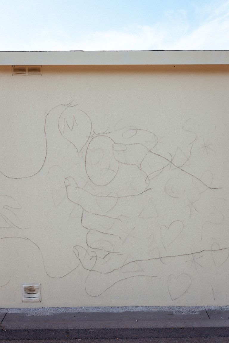 Faint pencil sketch outline of figures drawn on a light beige exterior building wall under a partly cloudy sky.