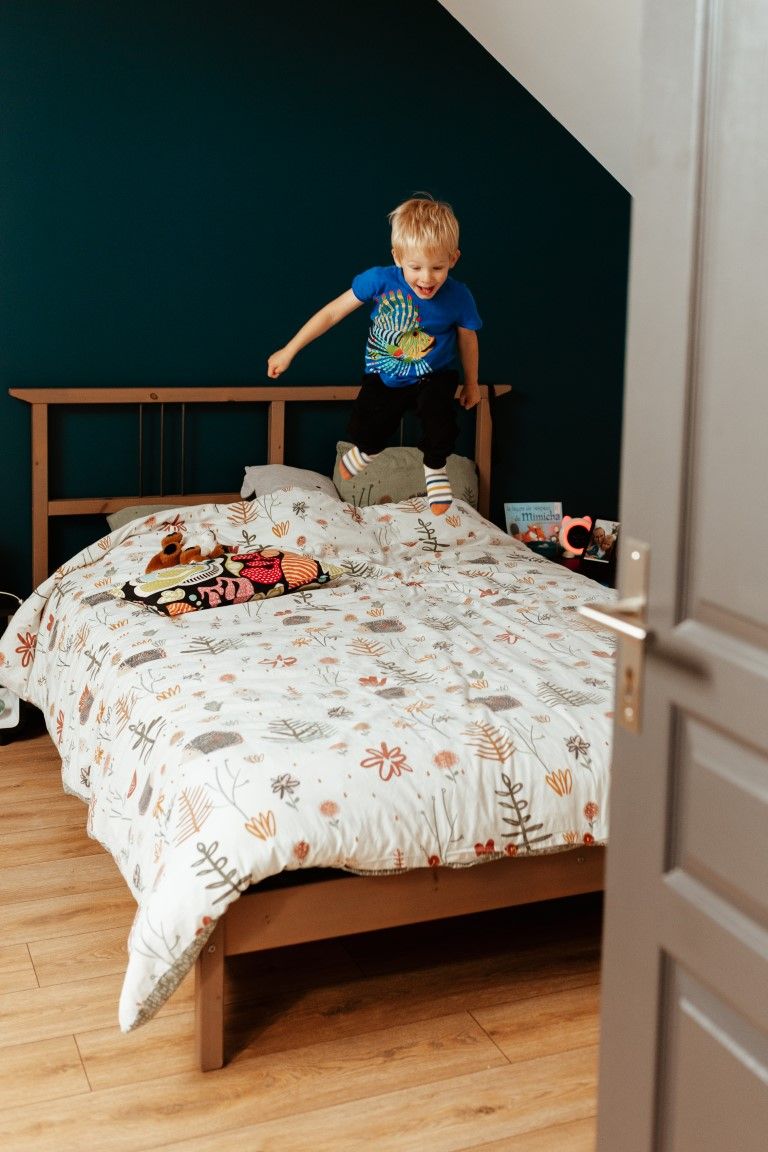 Un jeune garçon blond saute sur un lit dans une chambre aux murs turquoise.