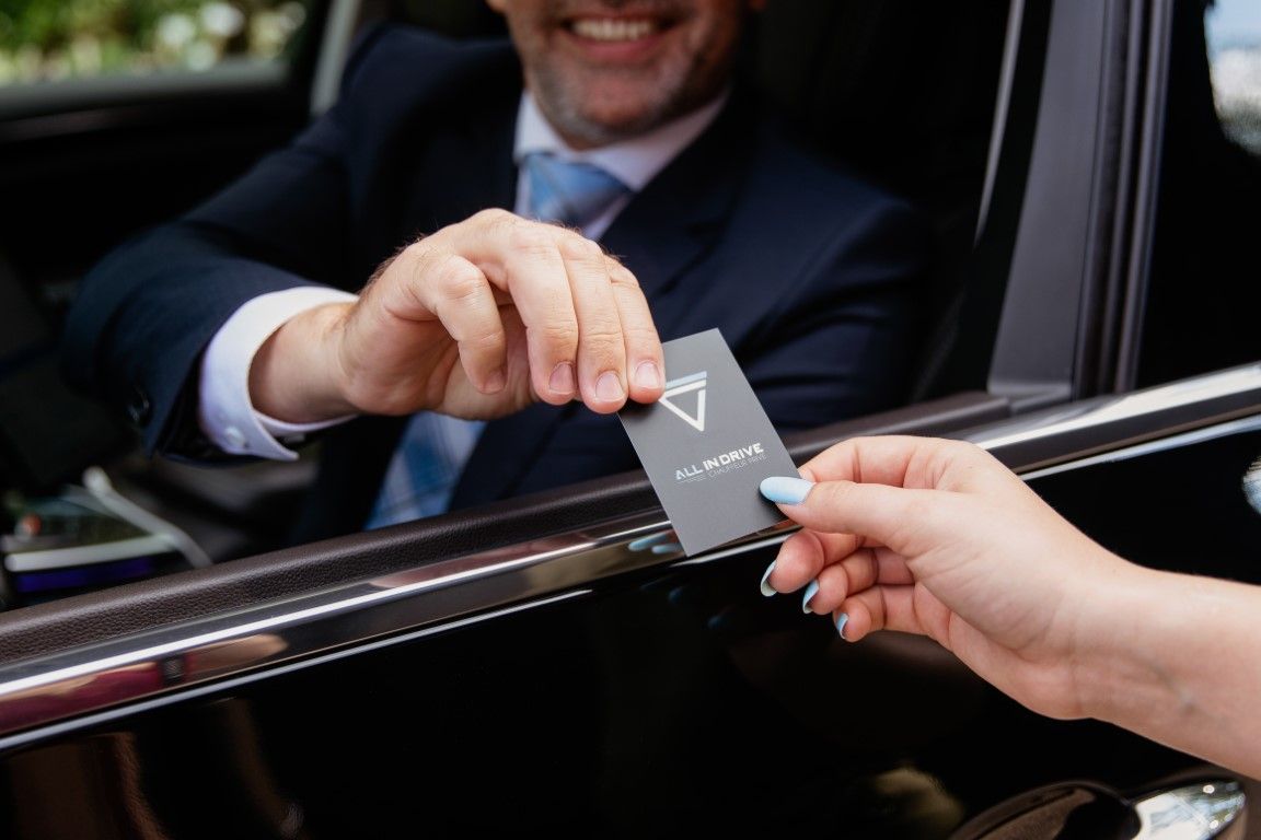 Smiling businessman in suit handing black business card through luxury car window.