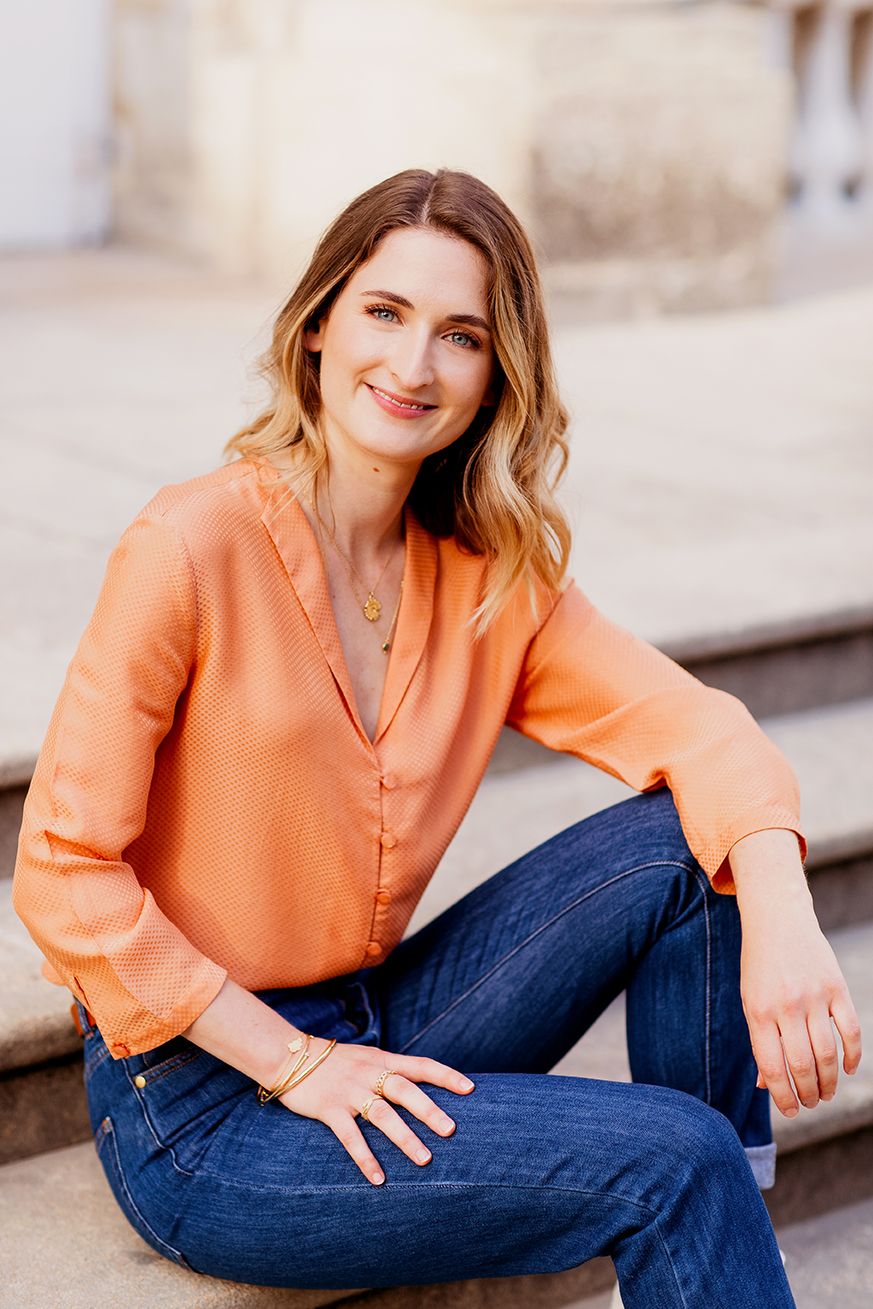 Woman in orange blouse and jeans sitting on steps, smiling in outdoor urban setting.