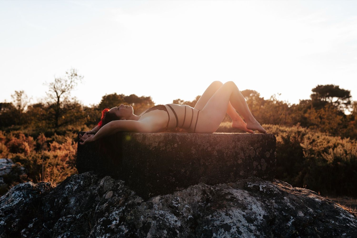 Person lying on a rock at sunset in a natural landscape with warm golden backlight and shrubs in the background.