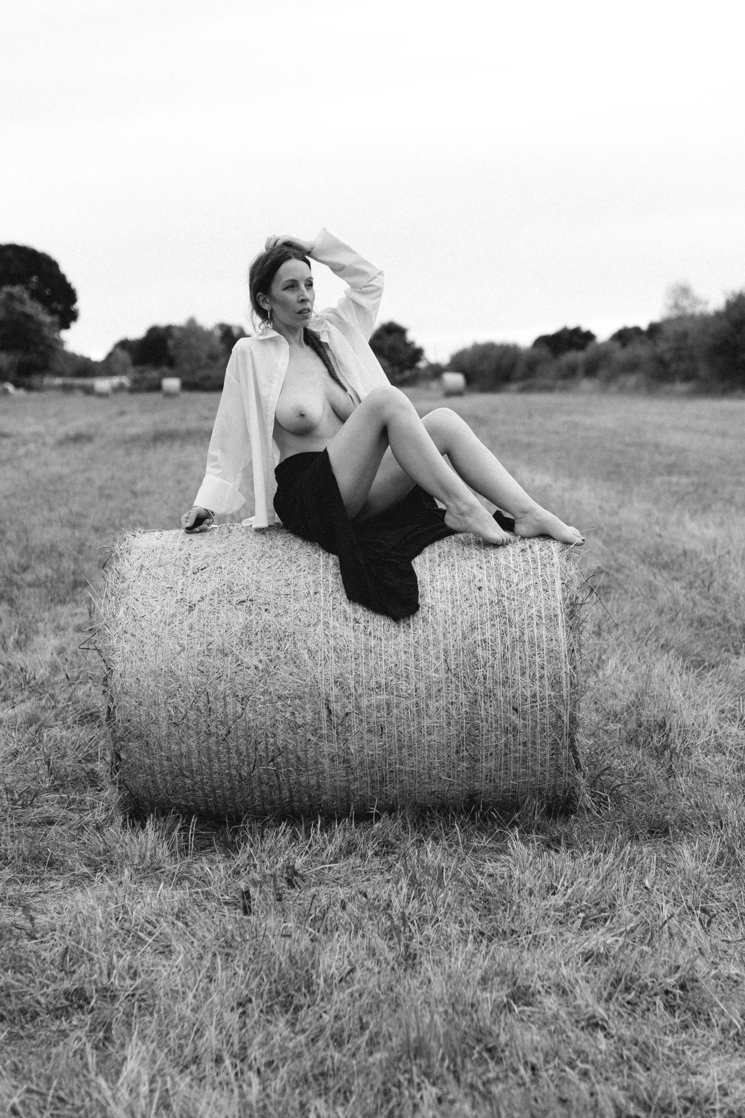 Black and white photo of a woman sitting on a hay bale in an open field, wearing an open white shirt and dark skirt.