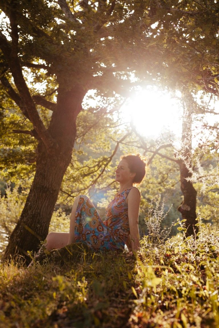 Woman in floral dress sitting under sunlit tree in golden hour light, surrounded by lush green grass and foliage.