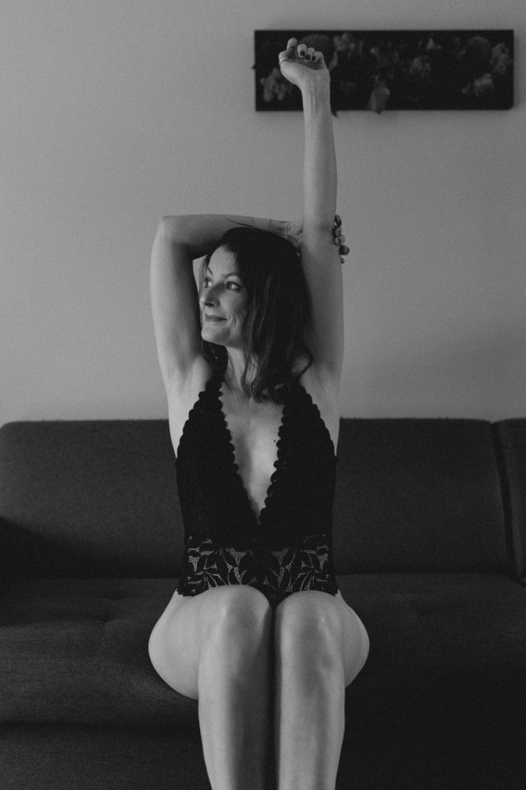 Photo en noir et blanc d'une femme en lingerie noire assise sur un canapé, les bras levés.