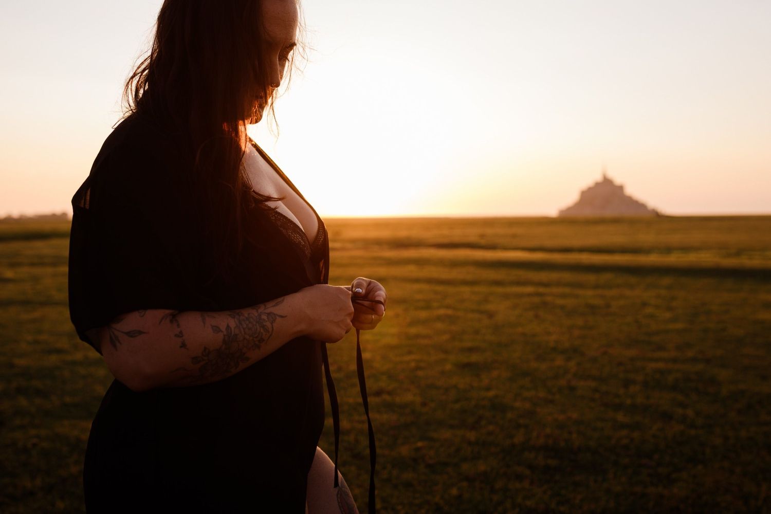 Woman with tattoo standing in silhouette against golden sunset with Mont Saint-Michel in background.
