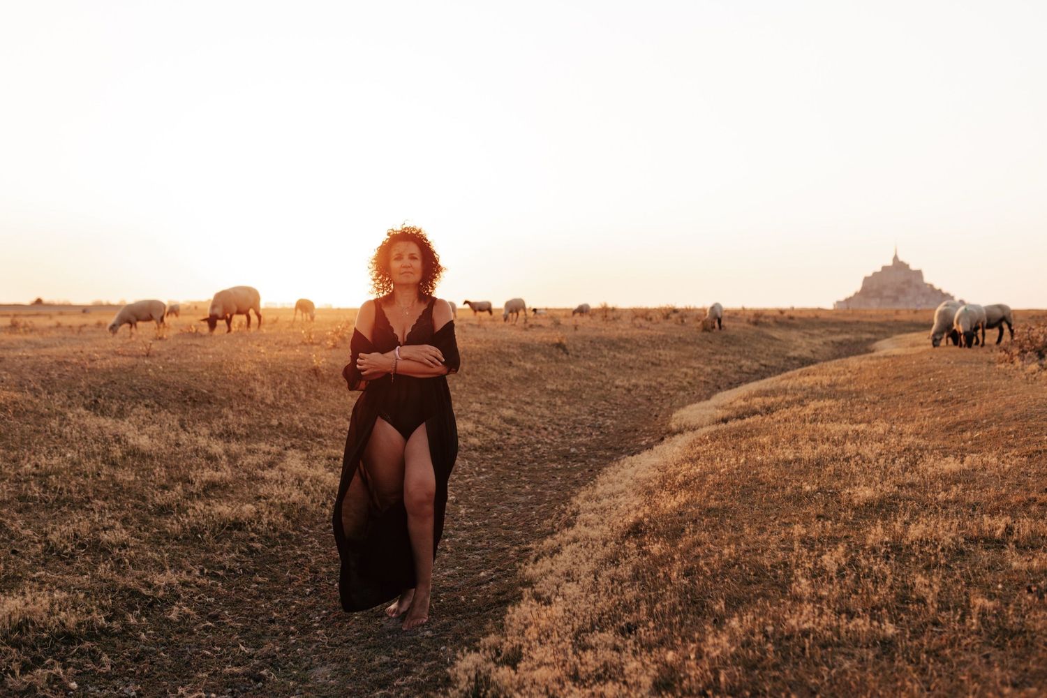 Woman in black dress walks through golden fields at sunset with horses grazing near Mont Saint-Michel.