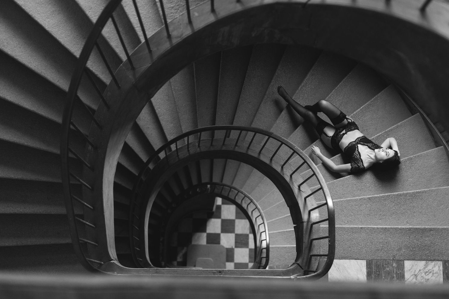 Black and white photo of a person lying on a spiral staircase, viewed from above, creating a dramatic geometric composition.