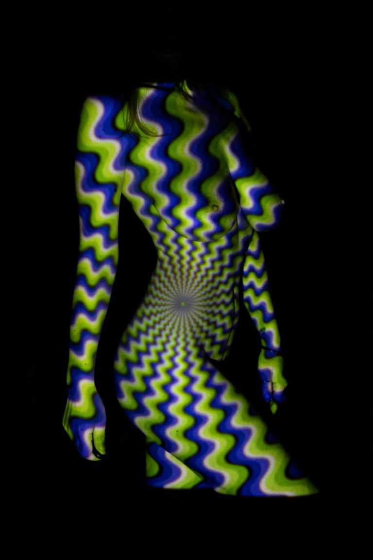 A human silhouette covered in vibrant green and blue hypnotic spiral wave pattern glowing against a black background.
