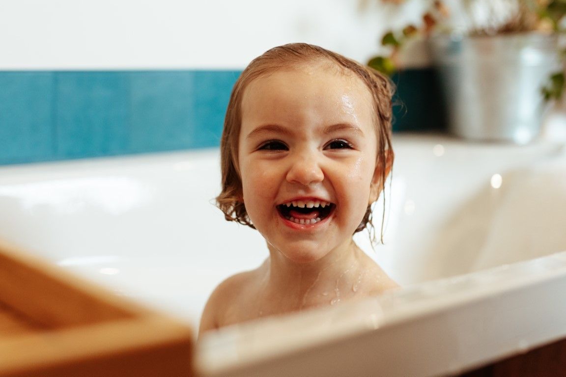 Un petit enfant riant aux éclats, les cheveux mouillés, assis dans une baignoire blanche.