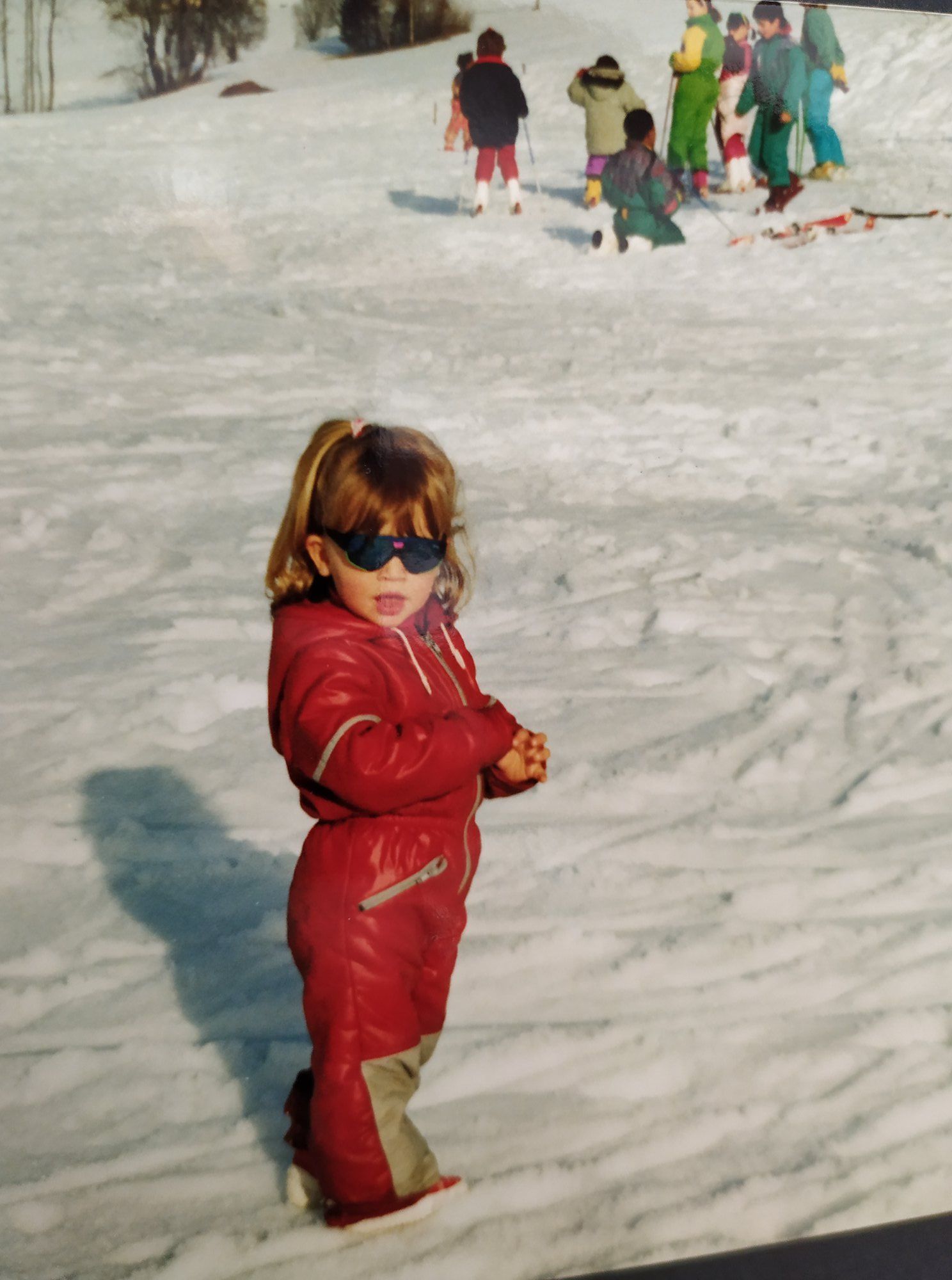 Jeune enfant en combinaison de ski rouge et lunettes de soleil posant sur une piste de ski enneigée.