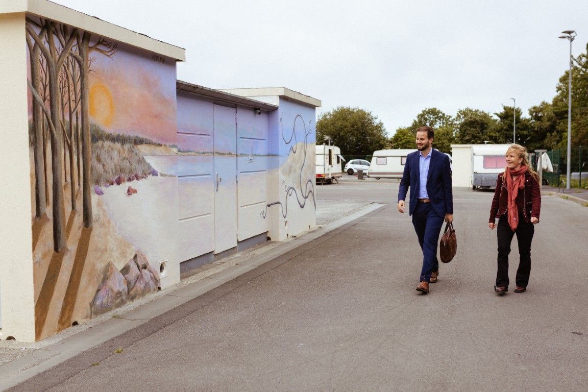 Two people walking past a colorful mural painted on a wall in an outdoor parking area on a sunny day.