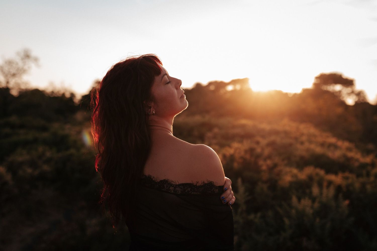 Woman with red hair basking in golden sunset light outdoors, wearing off-shoulder top in a serene nature setting.