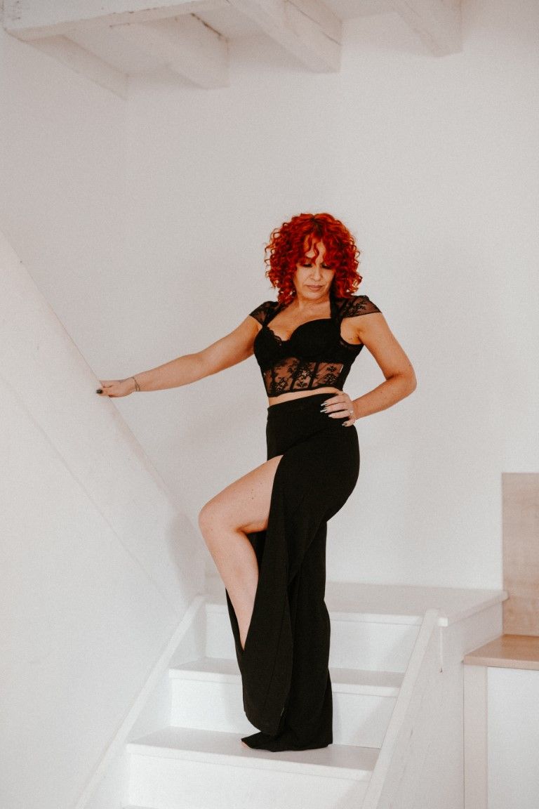 Woman with bold red curly hair wearing black crop top and high-slit maxi skirt on stairs.
