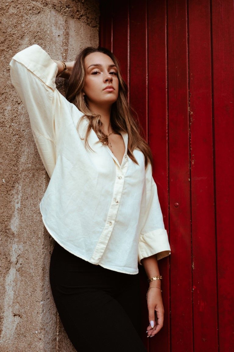 Jeune femme en chemise blanche boutonnée et pantalon noir posant devant une porte en bois rouge et un mur en pierre.
