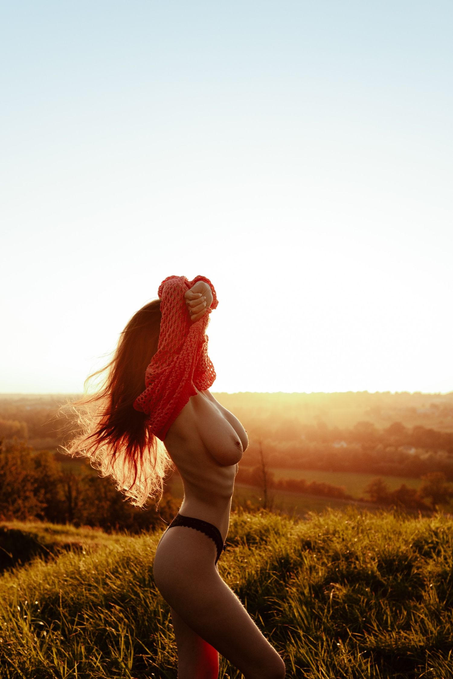 Woman holding red knit sweater outdoors at golden hour sunset with scenic countryside landscape in background.