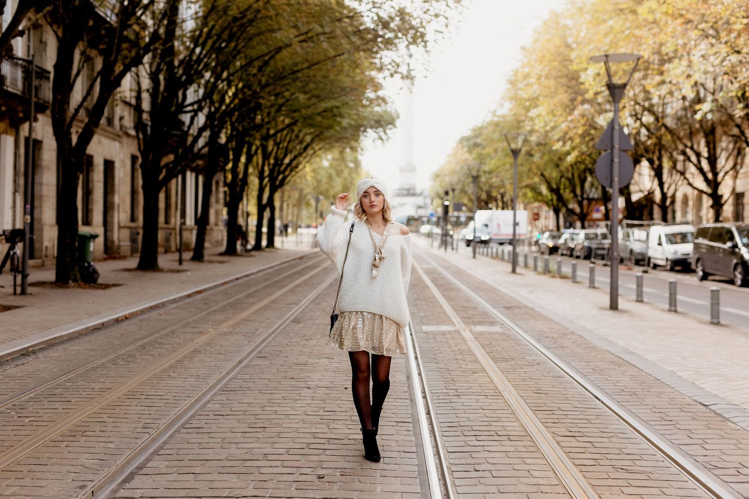 Une femme blonde en pull blanc et jupe courte marche sur les voies de tramway bordées d'arbres d'automne en ville.