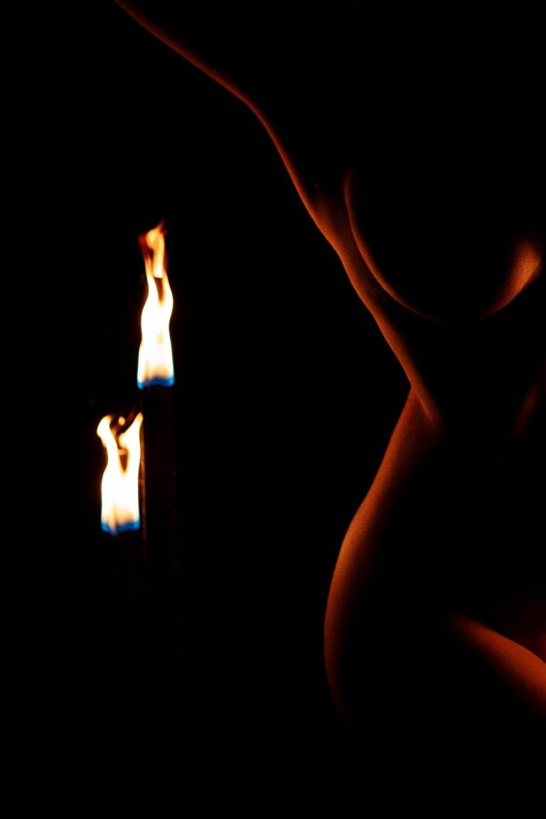 Artistic nude silhouette illuminated by two candle flames against a dark black background.