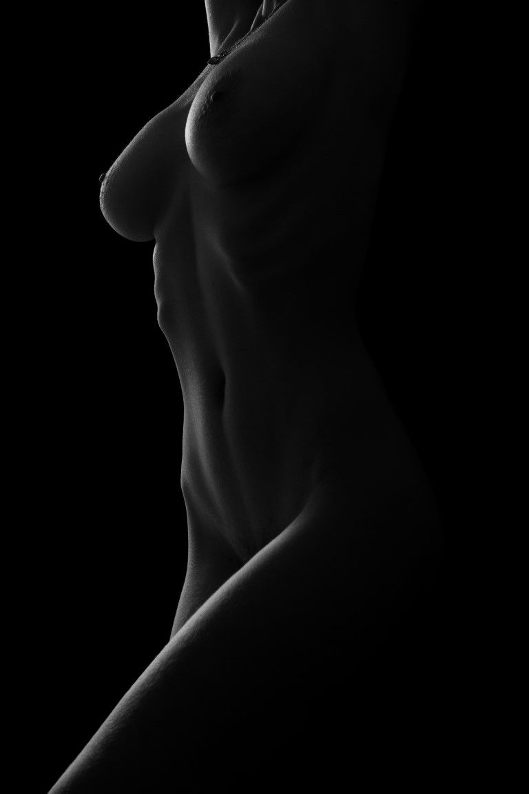 Black and white artistic nude photograph highlighting toned female torso with dramatic chiaroscuro lighting.