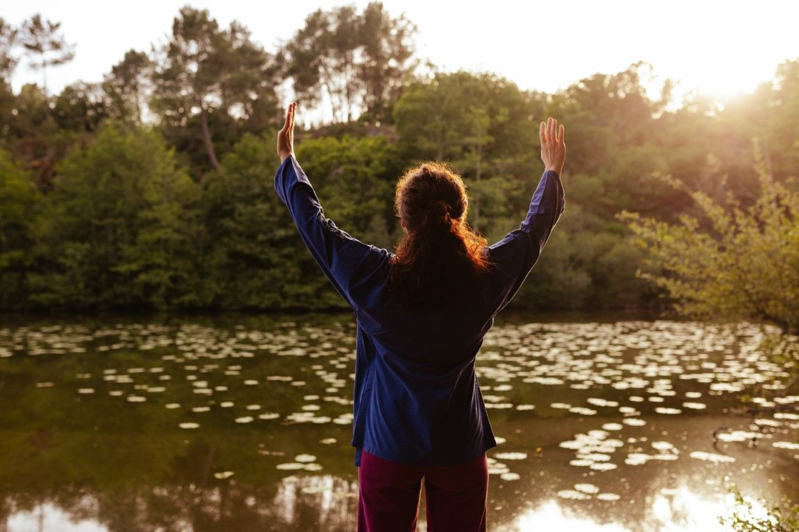Woman standing with arms raised facing a serene pond surrounded by lush green trees in golden sunlight.