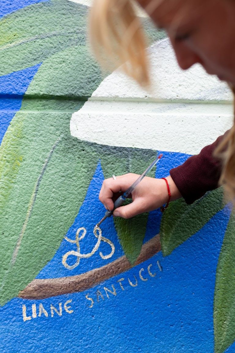 Artist Liane Santucci signing her name on a colorful blue and green painted mural outdoors.