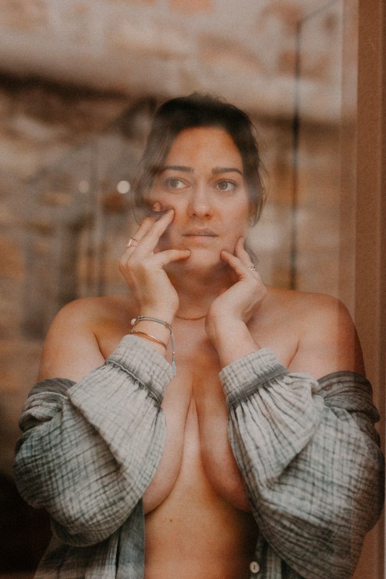 Woman in open grey shirt looking through glass window with hands framing her face.
