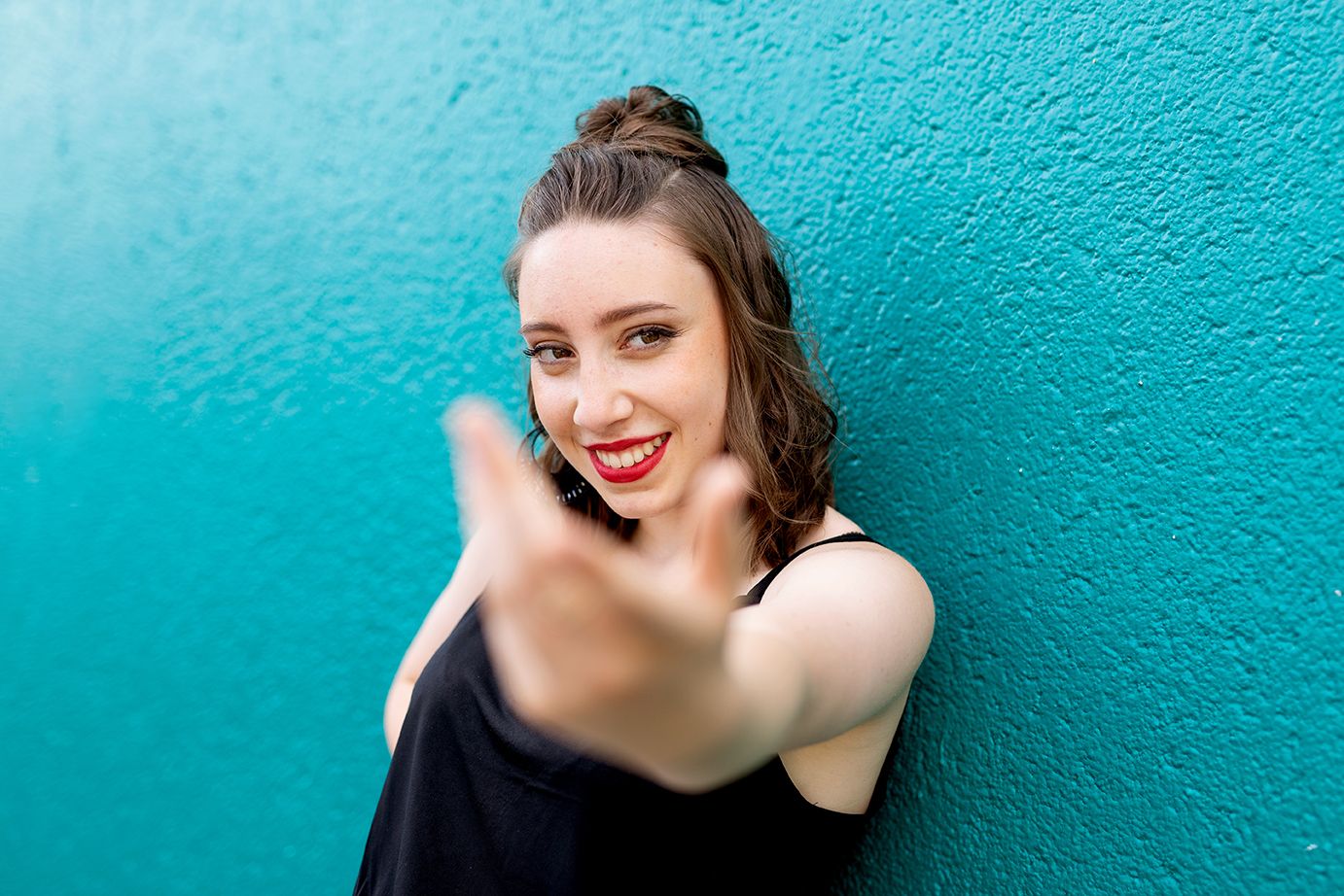 Femme brune souriante en haut noir tendant la main vers la caméra devant un mur turquoise en Basse Normandie.