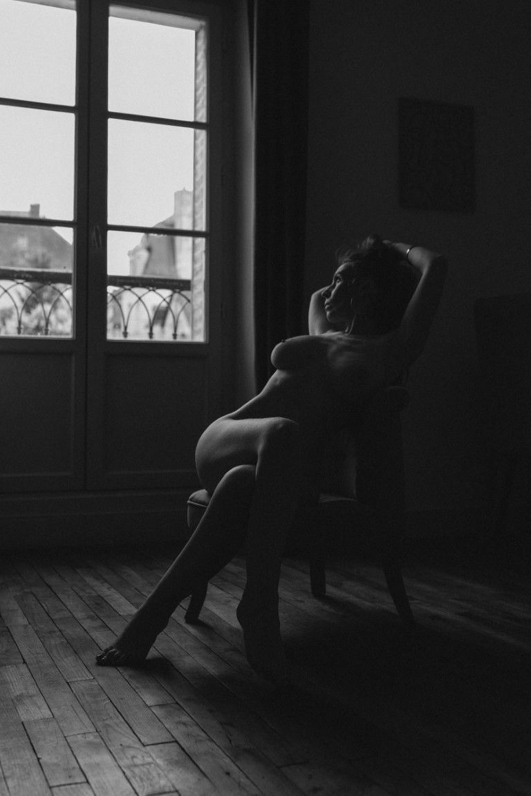 Silhouette en noir et blanc d'une femme nue allongée dans un fauteuil près d'une grande fenêtre, dans une lumière tamisée.