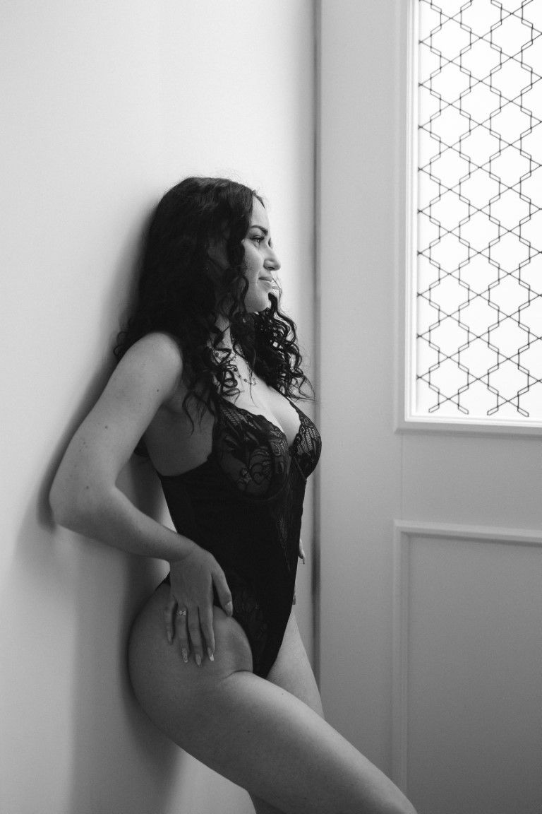Woman in black lace bodysuit leaning against white wall.
