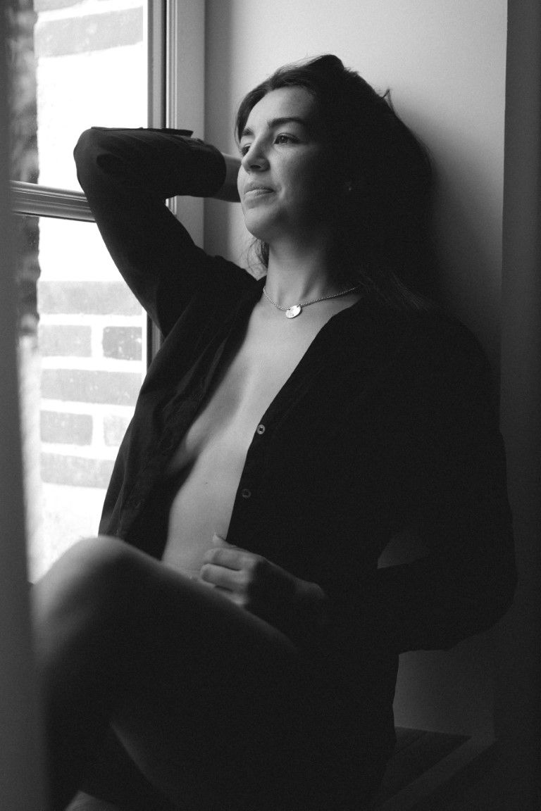 Black and white portrait of woman in open shirt sitting by window.