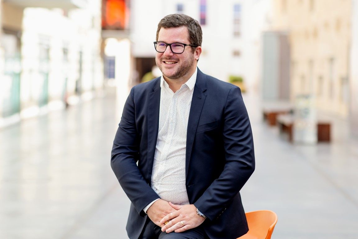 Smiling young man in blazer seated casually in modern bright public space.