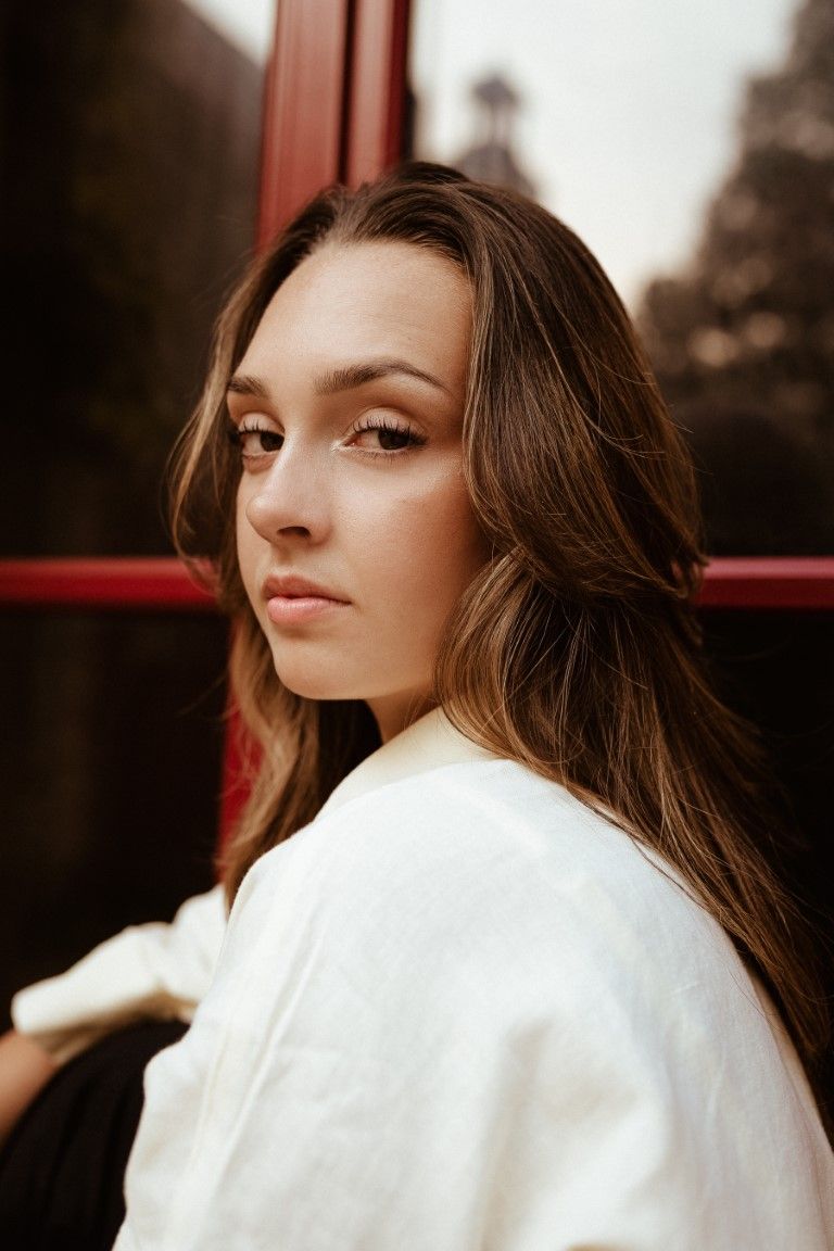 Jeune femme brune en veste blanche posant près d'une fenêtre à cadre rouge dans un décor urbain extérieur à l'atmosphère mélancolique.