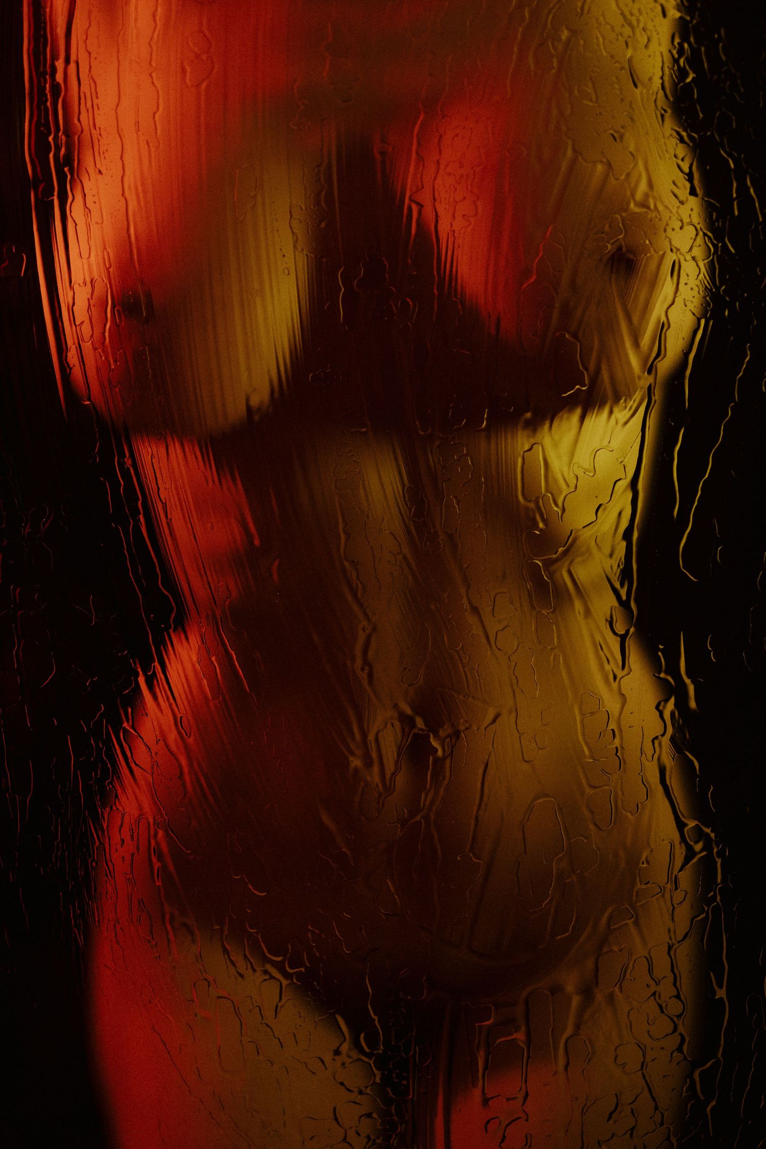 Artistic nude silhouette of a female figure seen through rain-streaked glass with warm red and gold lighting.