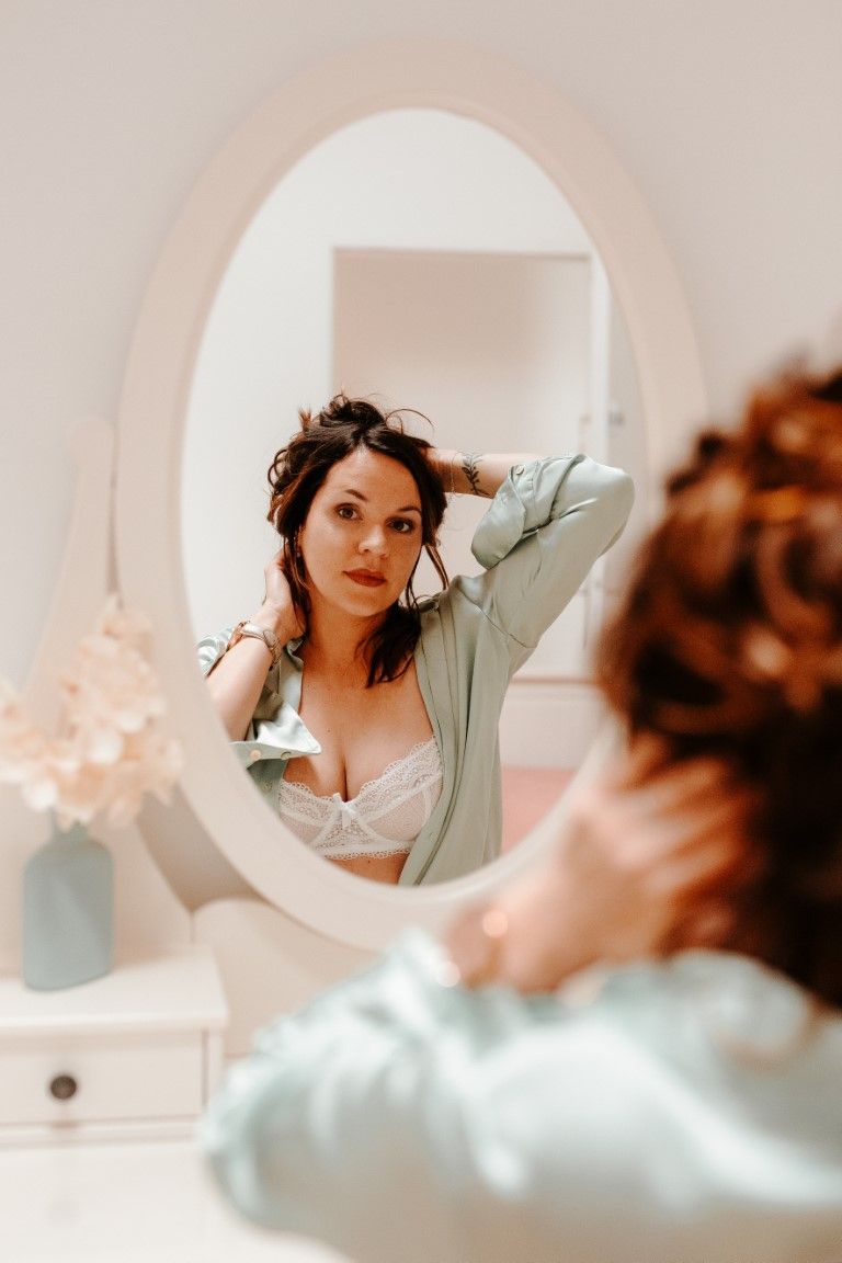 Woman in white lace bra and blue robe fixing her dark hair while looking at her reflection in an oval mirror.