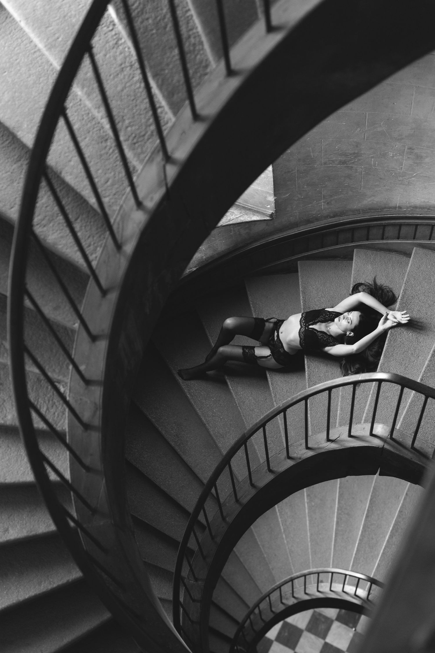 Black and white aerial view of a person lying on a grand spiral staircase, creating a dramatic architectural composition.