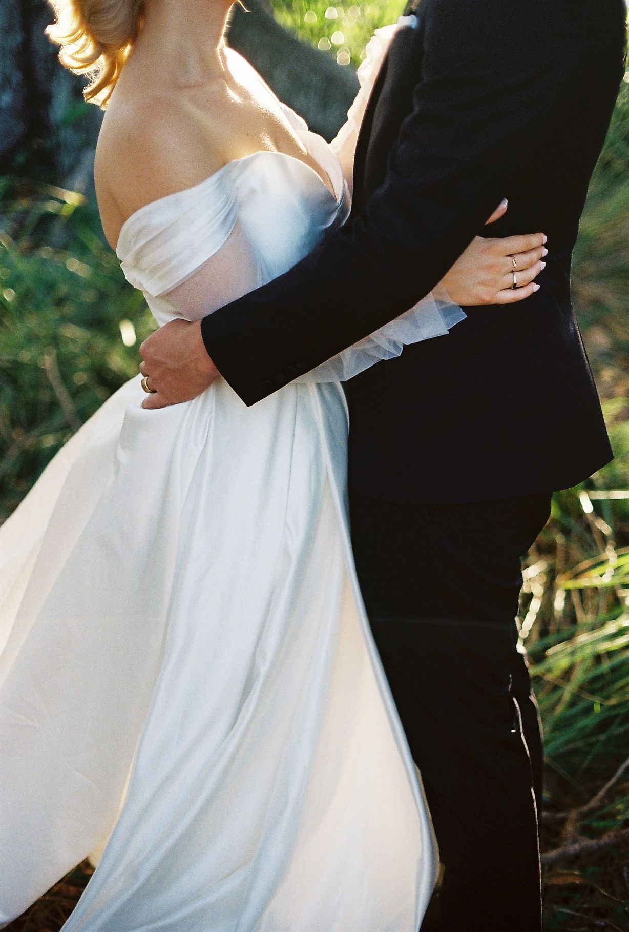 Close-up of bride in off-shoulder white gown and groom in black tuxedo embracing outdoors in warm sunlight.