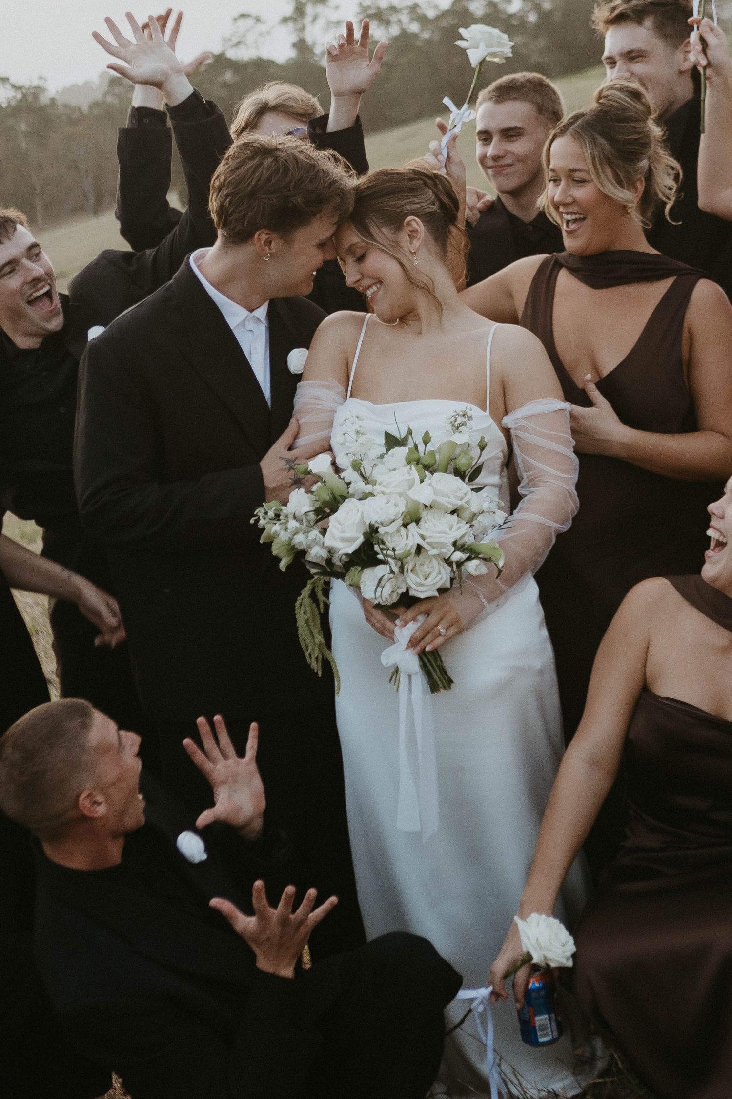 Bride and groom share an intimate moment surrounded by cheering wedding party outdoors, bride holding white floral bouquet.