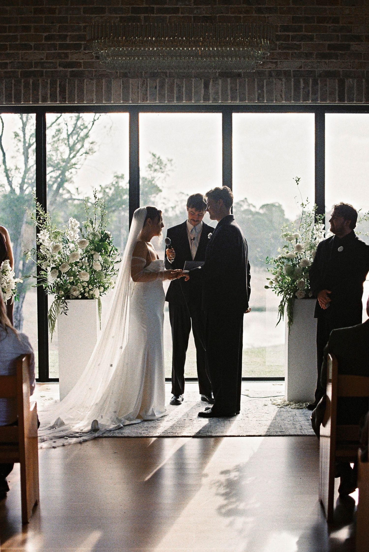 Bride and groom exchange vows at elegant indoor wedding ceremony with white floral arrangements and large windows.