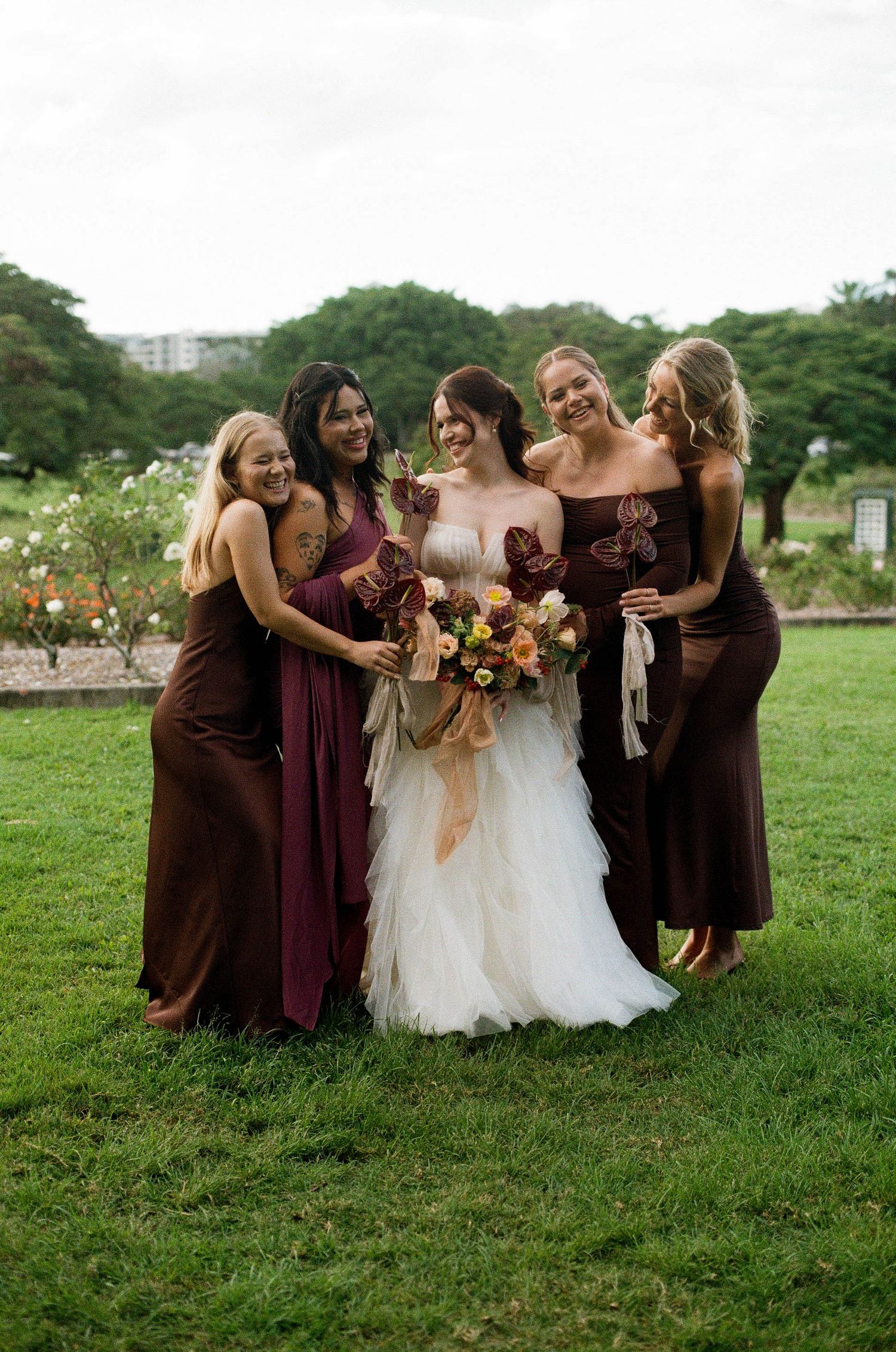 Bride in white tulle gown smiles with four bridesmaids in brown dresses holding bouquets on green lawn.