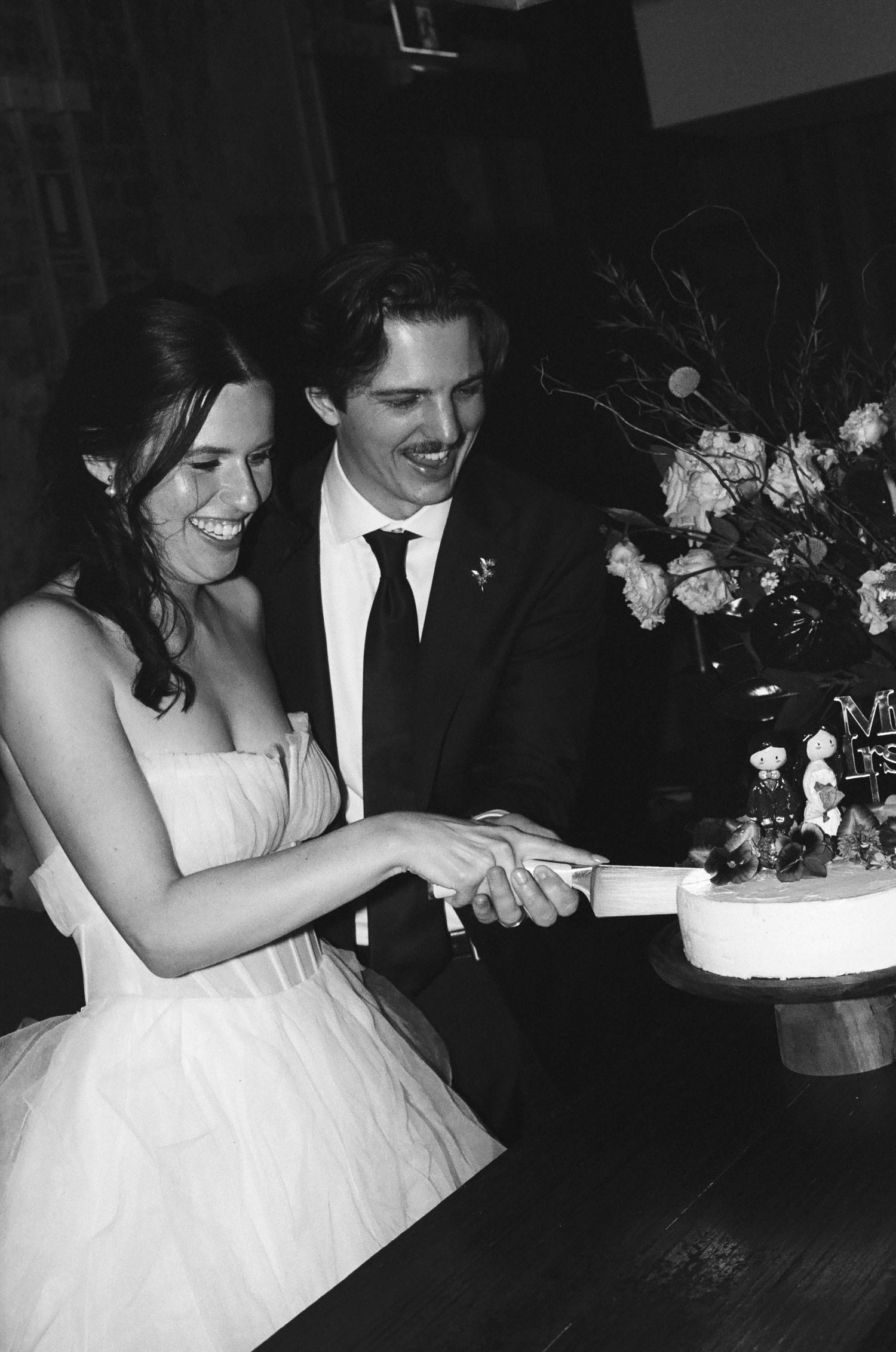 Smiling bride and groom cutting their wedding cake together at their reception in black and white photo.