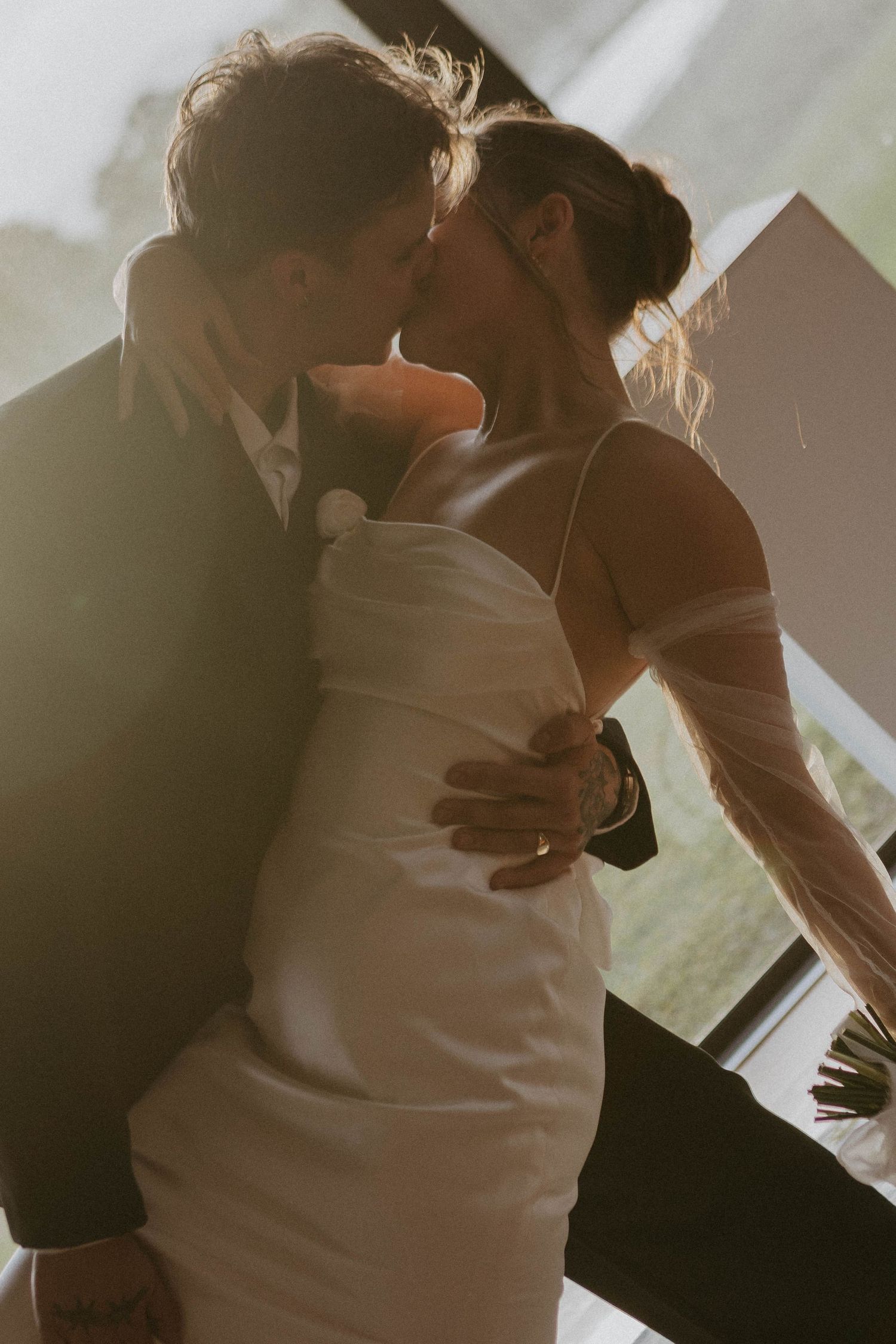 A bride and groom share a romantic kiss in golden sunlight, the bride wearing a white satin gown with sheer gloves.
