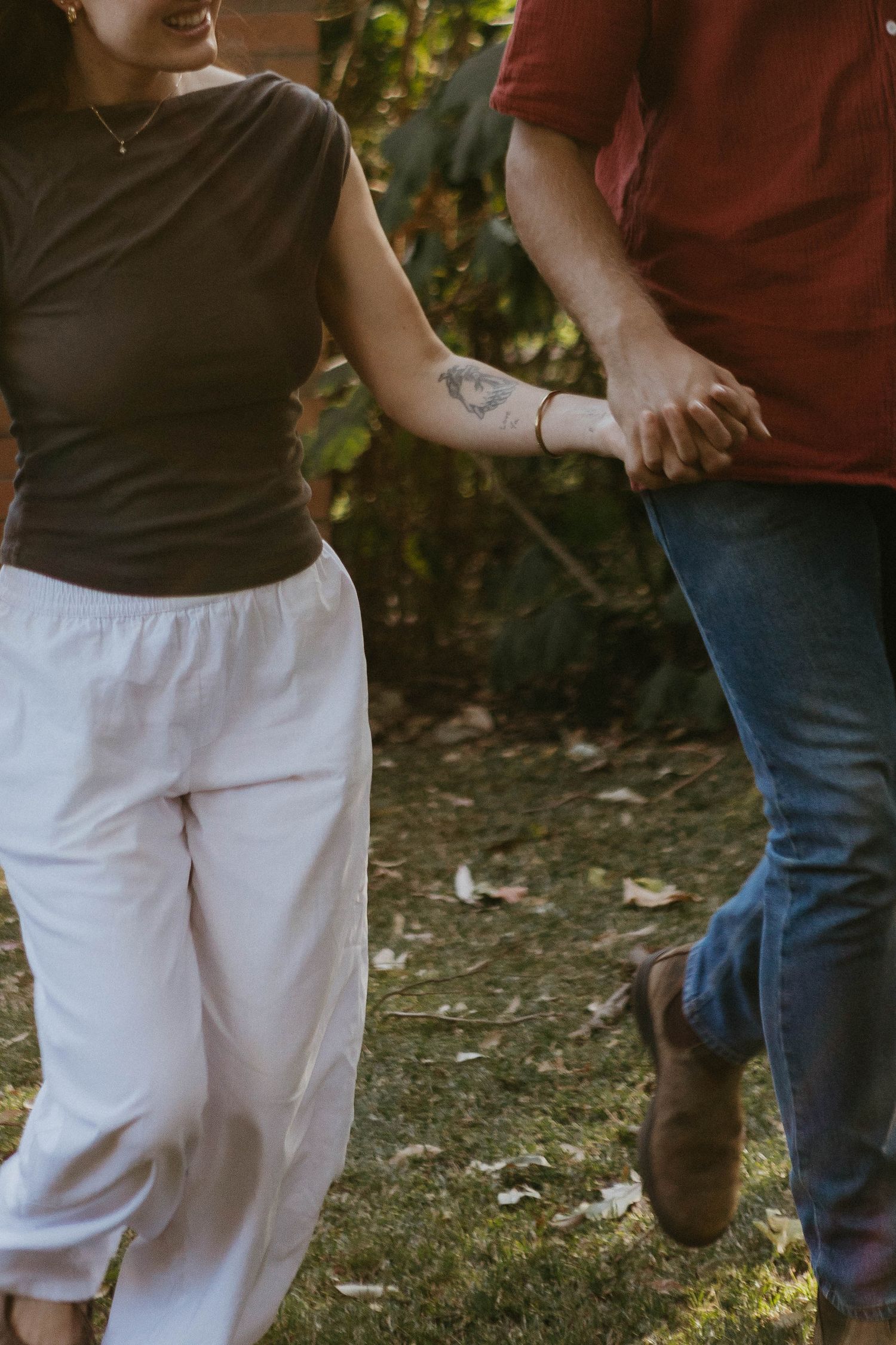A couple holds hands while walking outdoors, the woman wearing white pants and a dark top with a floral tattoo on her arm.