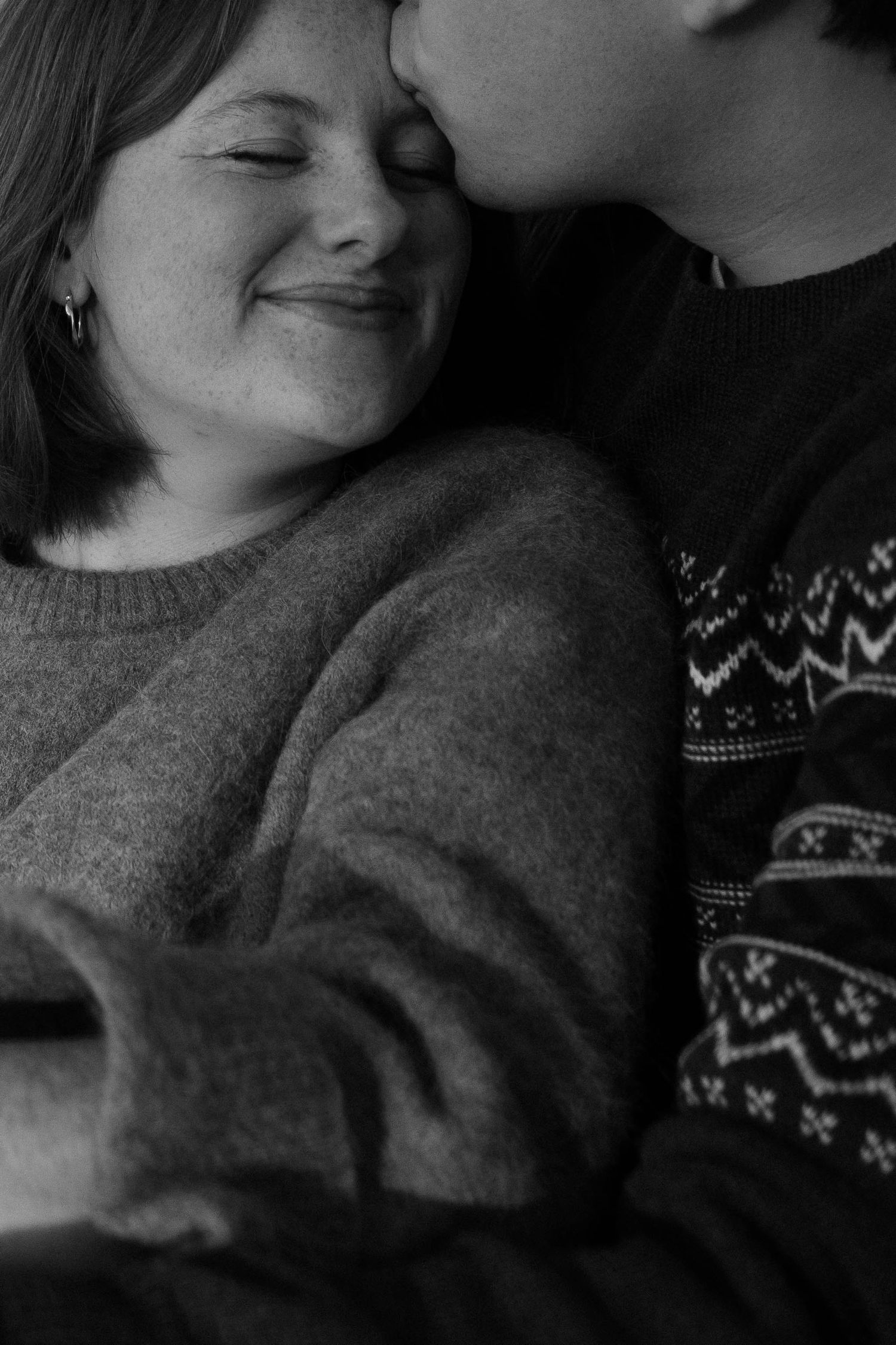Black and white photo of a smiling woman being kissed on the cheek by a man in a patterned sweater.