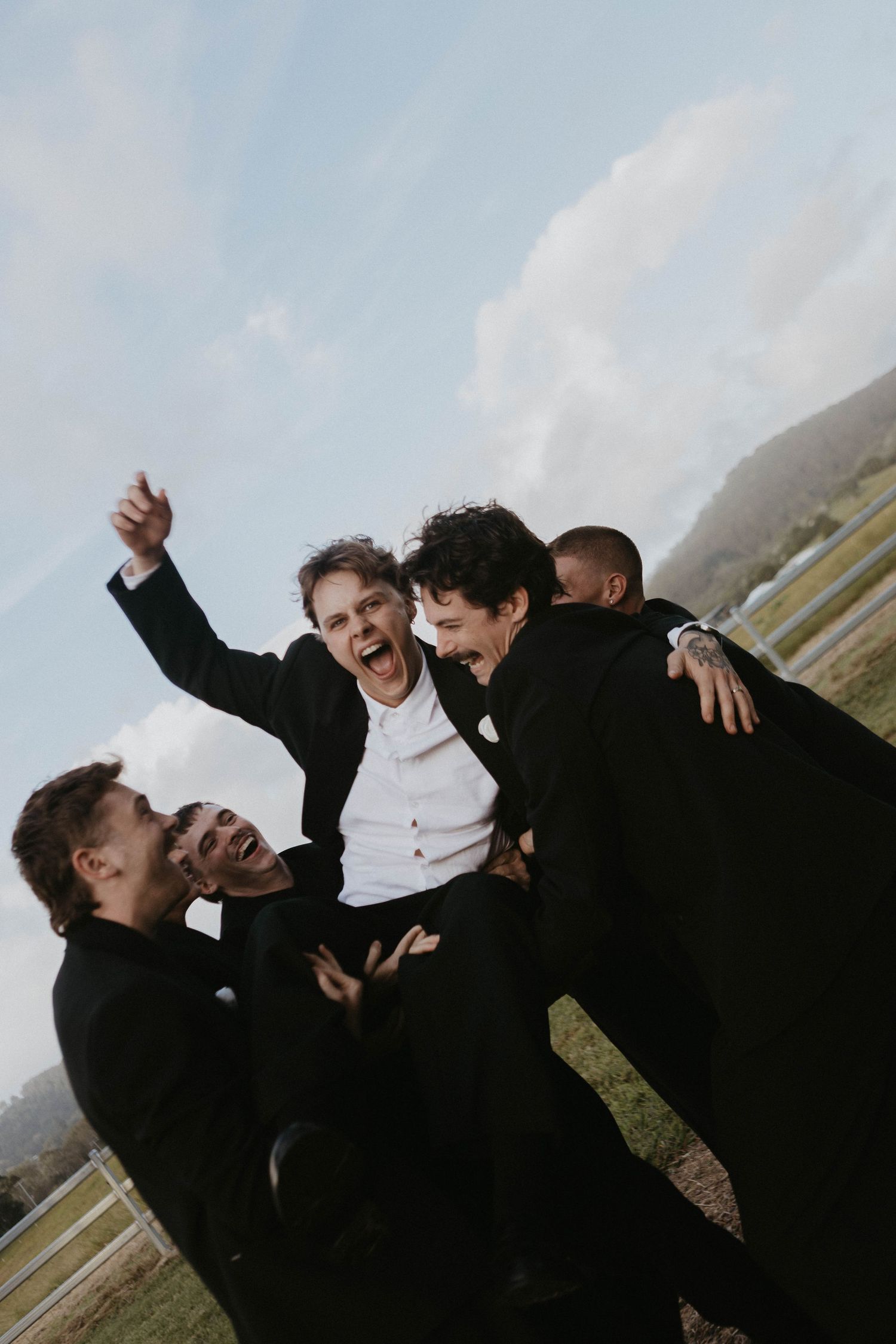 Groomsmen in black suits celebrating joyfully outdoors, lifting the groom with laughter and cheers on a wedding day.