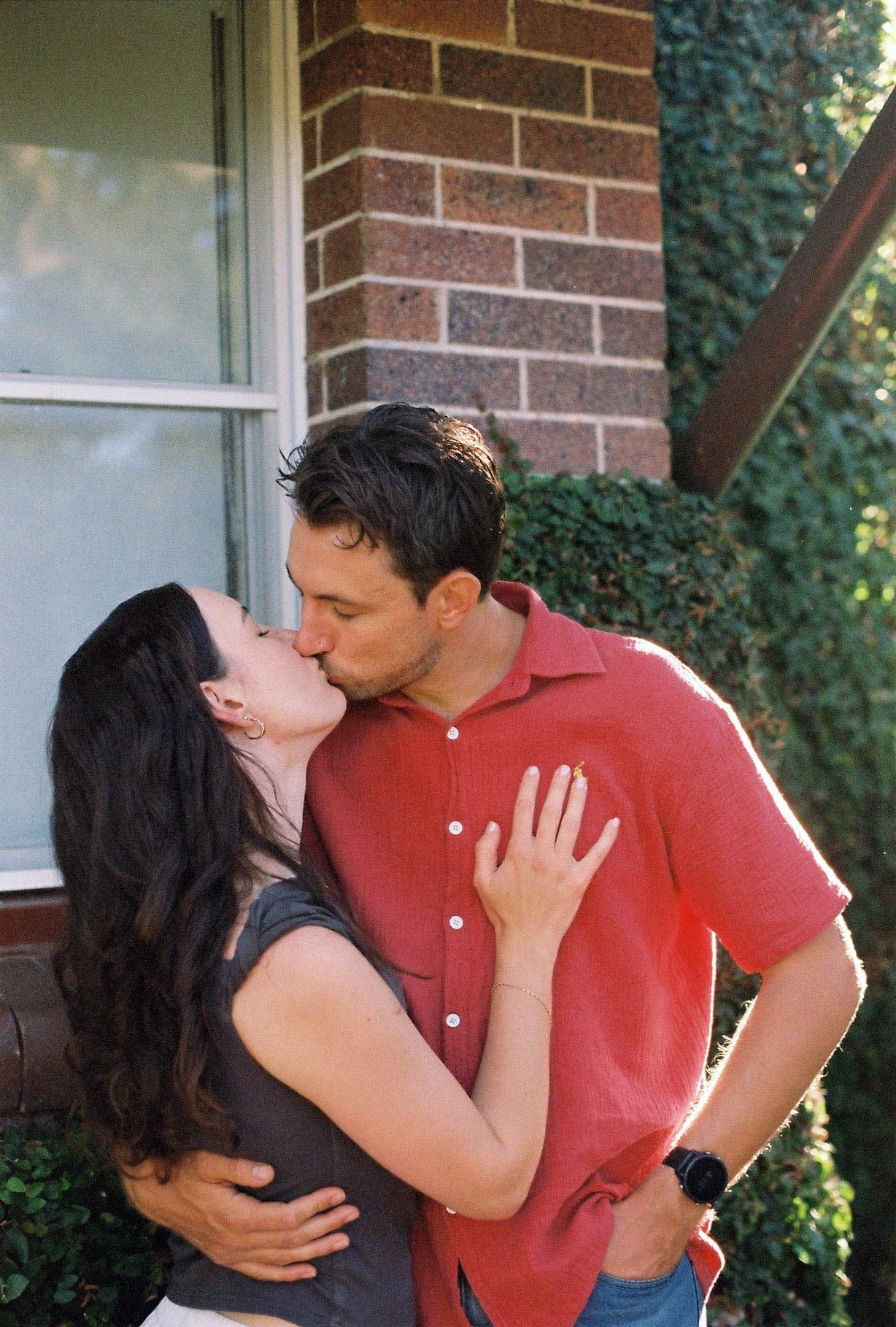 A couple sharing a romantic kiss outside a brick building with ivy, the woman in gray and man in red shirt.